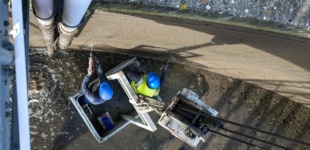 Zwei Mitarbeiter in Schutzkleidung und blauen Helmen sind von oben zu sehen. Die bohren in die Innenwand des Klärwerks.