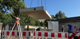 Baustelle mit schwebender Betonplatte, rot-weißen Absperrgittern, Vermessungsgerät, Arbeitern und Bäumen im Hintergrund.